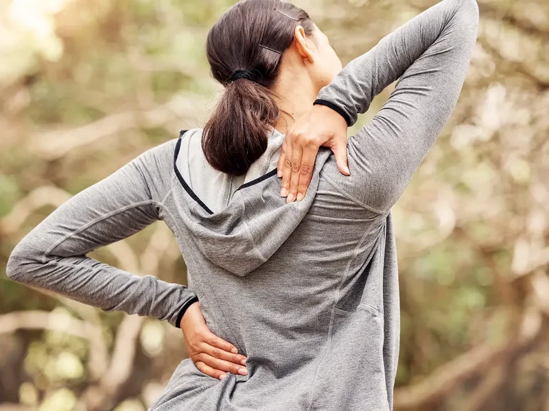 Woman outdoors reaching one hand behind her back and the other over her shoulder while stretching for lower back pain